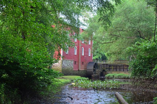 A red mill building beside a calm stream, featuring a water wheel and surrounded by lush green trees and foliage.