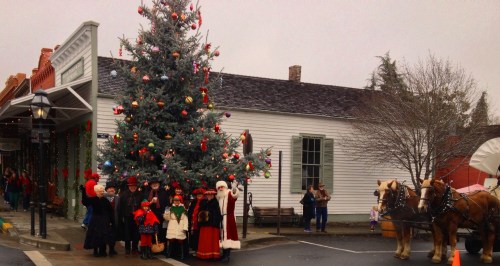 A festive scene with a large Christmas tree, people in holiday attire, and a horse-drawn carriage in a quaint town.