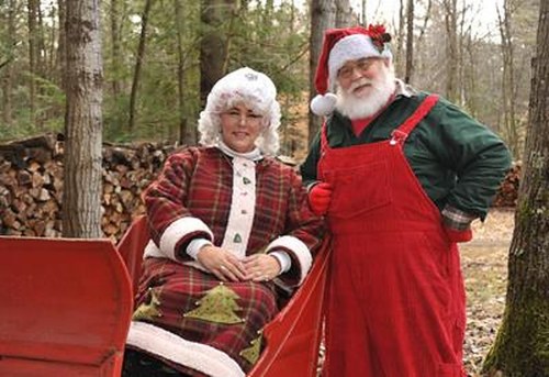 A man in a Santa outfit and a woman in a festive dress sit in a red sleigh surrounded by trees and firewood.