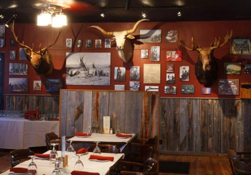 Rustic restaurant interior with wooden walls, red accents, and mounted animal heads, featuring tables set for dining.