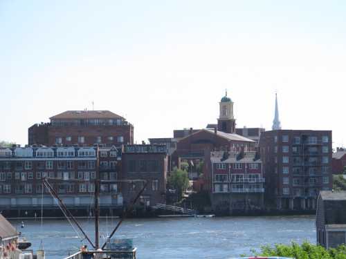 View of a waterfront cityscape featuring brick buildings and a church steeple against a clear blue sky.