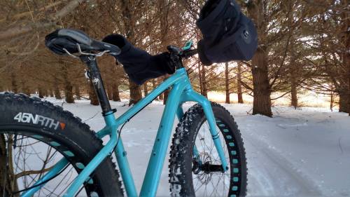 A turquoise fat bike parked on a snowy trail surrounded by trees, with a black bag attached to the handlebars.