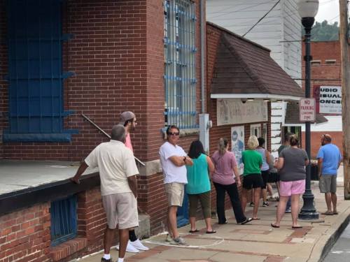 A line of people waiting outside a brick building on a sidewalk, with some standing and others sitting.