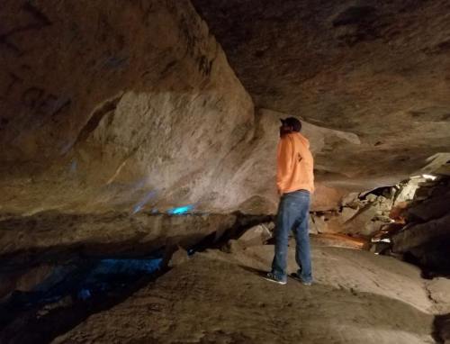 A person in an orange hoodie stands in a dimly lit cave, gazing at the rock formations and shadows around them.