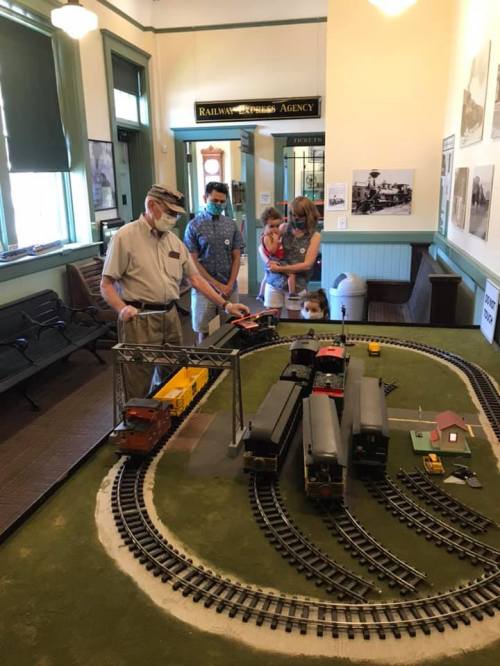 An elderly man operates a model train set while a family observes in a historical railway station.