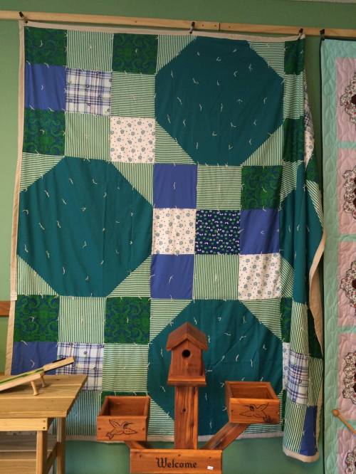 A colorful quilt featuring green, blue, and patterned squares and hexagons, displayed on a wall.