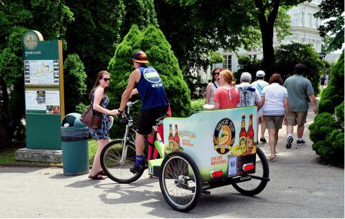 A cyclist with a colorful cart sells drinks to people walking in a park, surrounded by greenery and a signpost.