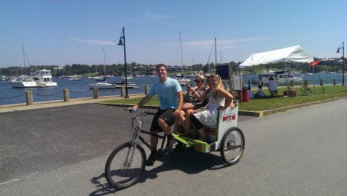 A group of four people enjoying a ride in a rickshaw by a marina with boats and a tent in the background.