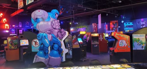 A vibrant arcade filled with classic game machines and a large pixelated character mural on the wall.