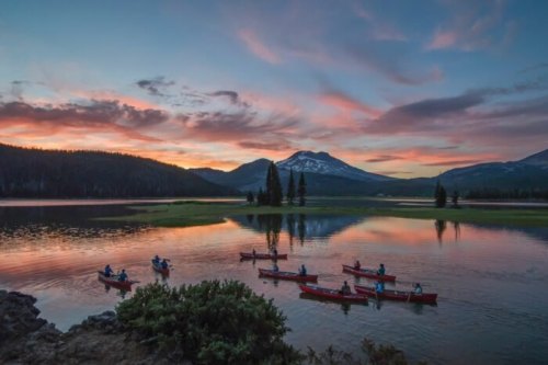 A serene lake at sunset with kayakers paddling, surrounded by mountains and colorful clouds reflecting on the water.