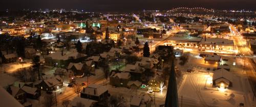 A nighttime aerial view of a snowy town, illuminated by colorful lights and a bridge in the background.