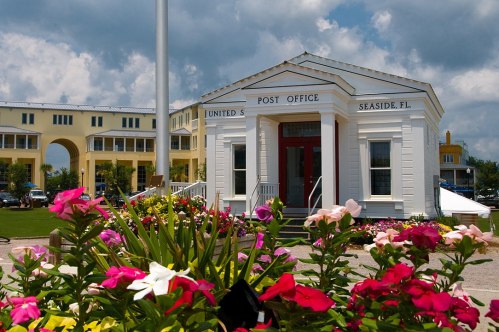 A charming white post office building surrounded by colorful flowers in Seaside, Florida, under a partly cloudy sky.