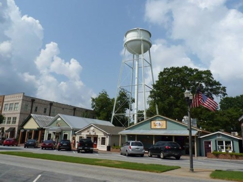 A small town scene featuring a water tower, shops, and parked cars under a partly cloudy sky.