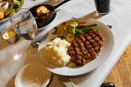 A plate with grilled steak, mashed potatoes, and sautéed vegetables, accompanied by a glass of water and a dark beverage.
