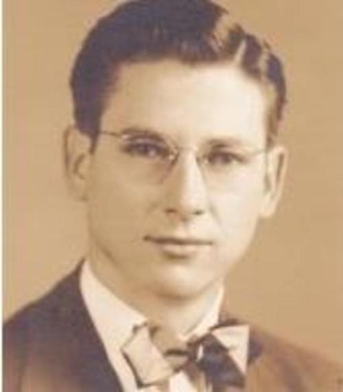 A young man in a suit and bow tie, wearing glasses, with a neutral expression against a sepia-toned background.