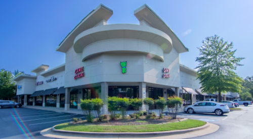 A modern retail plaza with multiple storefronts and a clear blue sky, featuring green landscaping and parked cars.