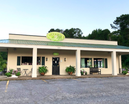 A charming restaurant exterior with a green sign, potted plants, and outdoor seating in a serene setting.