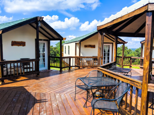A wooden deck with tables and chairs, overlooking green fields and blue skies, featuring two cozy cabins.