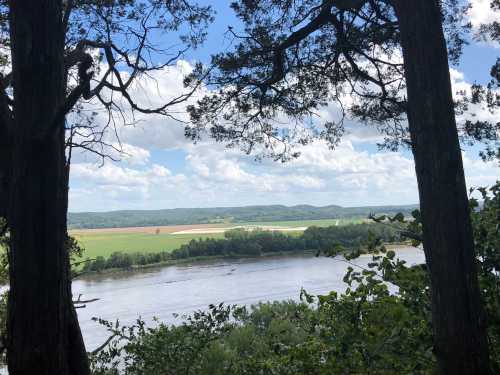 A scenic view of a river surrounded by lush greenery and fields under a partly cloudy sky.