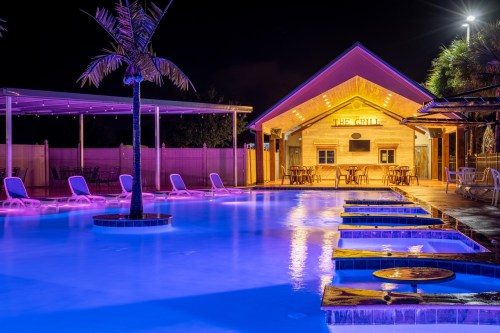 A vibrant pool area at night, featuring colorful lights, lounge chairs, and a grill house in the background.