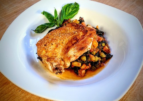 A plate featuring a golden-brown roasted chicken thigh on a bed of mixed vegetables, garnished with fresh basil.