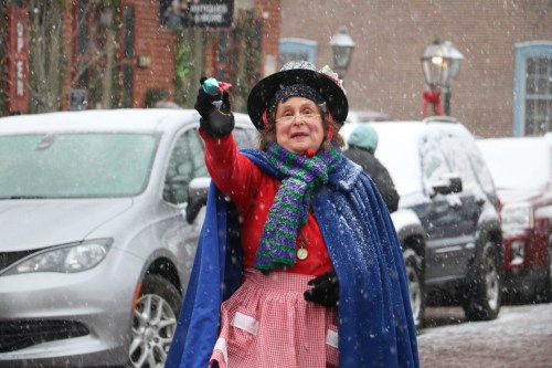 A cheerful performer in a colorful outfit and hat holds a toy while snow falls around her in a festive street scene.