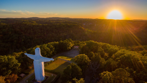 Aerial view of a large statue of Christ at sunset, surrounded by lush green trees and hills.