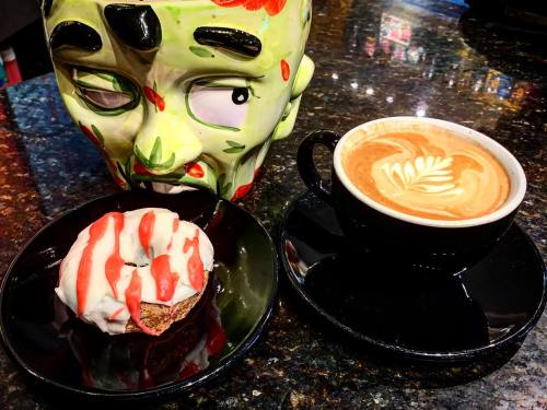 A colorful zombie mask beside a donut with icing and a latte with latte art on a black plate and cup.