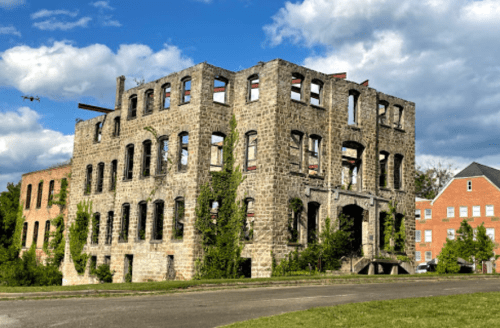 Abandoned stone building with large windows, partially covered in greenery, set against a cloudy sky.