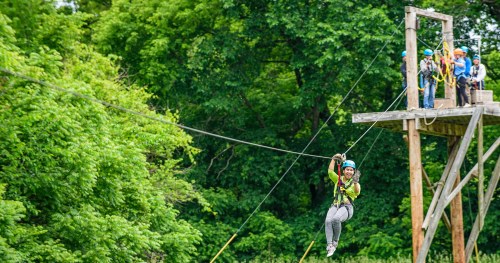 A person zip-lining through lush greenery, with a group of people on a platform in the background.