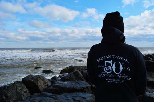 Person in a black hoodie stands on rocky shore, facing turbulent ocean waves under a cloudy sky.
