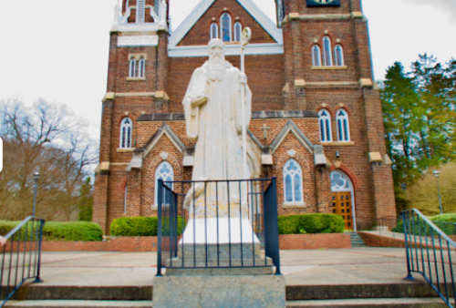 A tall white statue of a figure stands in front of a brick church with large windows and greenery surrounding it.