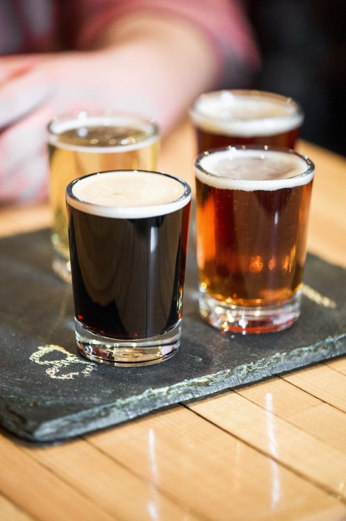 A slate tasting board with four small glasses of different beers: a dark stout, a light lager, and two amber ales.