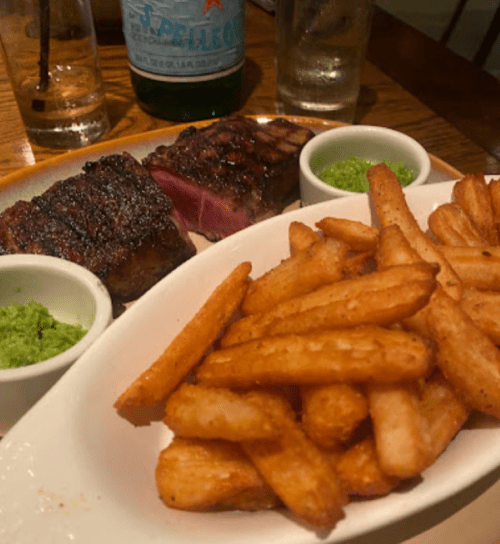 A plate of crispy fries with two cuts of steak and green sauce in small bowls, set on a wooden table.