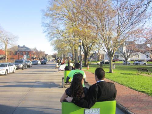 A couple enjoys a ride in a green pedicab on a tree-lined street, with other pedicabs and cars in the background.