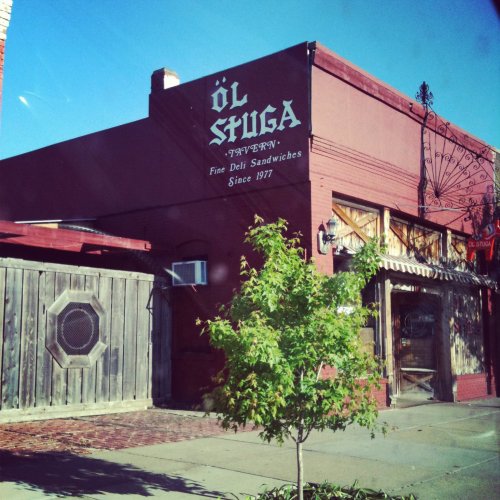 A rustic deli building with a sign reading "ÖL STUGA" and greenery in front, under a clear blue sky.