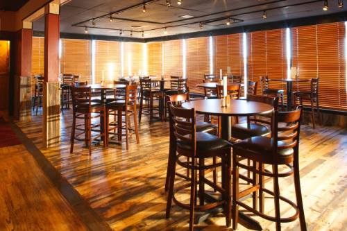 A warmly lit restaurant interior with wooden floors, empty tables, and blinds partially drawn on large windows.