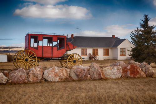 A red stagecoach sits on a grassy area in front of a white building and rocky landscape under a blue sky.