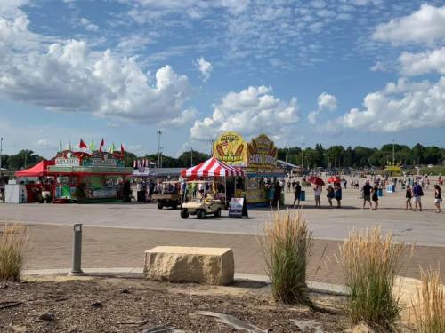 A lively fair scene with food stalls, a golf cart, and crowds under a blue sky with fluffy clouds.