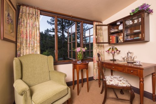 Cozy room with a green armchair, wooden table, and window showcasing trees outside, decorated with floral curtains.