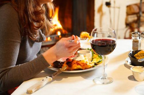 A person enjoying a meal by a fireplace, with a glass of red wine and a plate of food on the table.