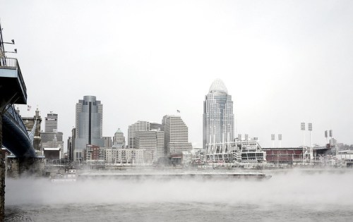 Foggy riverfront view of a city skyline with tall buildings and a baseball stadium in the background.