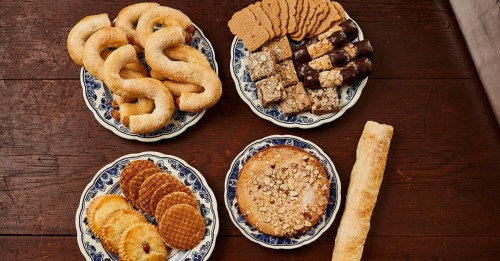 A variety of baked goods on decorative plates, including cookies, bars, and pastries, arranged on a wooden table.