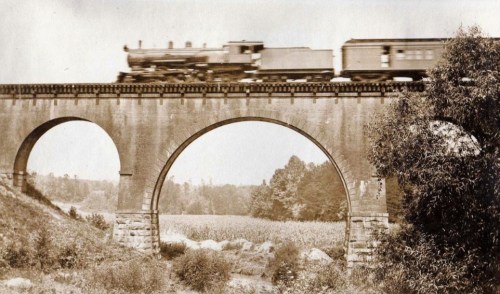 A vintage black-and-white photo of a train crossing a stone bridge over a rural landscape.