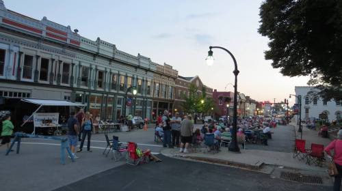 A lively street scene at dusk with people gathered in chairs, enjoying an outdoor event in a small town.