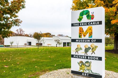 Sign for The Eric Carle Museum of Picture Book Art, featuring colorful illustrations and a modern building in the background.