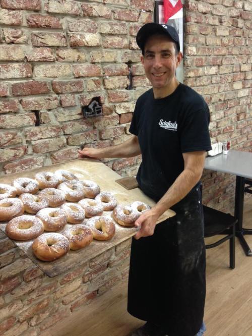 A smiling baker holds a tray of freshly baked pastries in a rustic brick bakery.