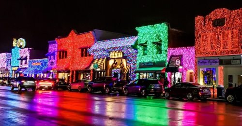 Colorful holiday lights adorn buildings along a street, reflecting on wet pavement at night.