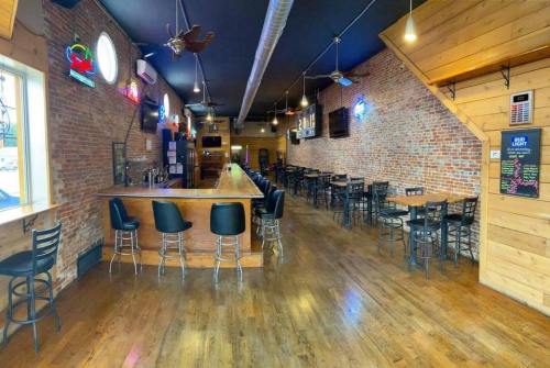 Interior of a bar with wooden floors, brick walls, and a long bar counter lined with black stools. Neon signs illuminate the space.