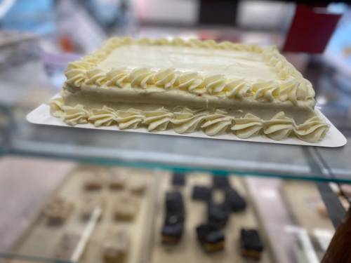 A rectangular cake with white frosting and decorative piping, displayed in a glass case with assorted desserts in the background.